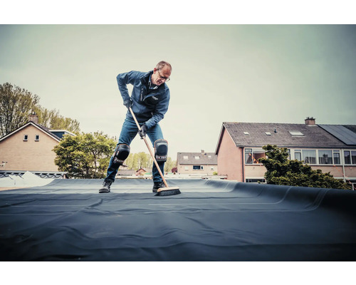 Een man veegt dakbedekking op een plat dak met een bezem.