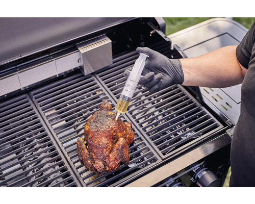 Een persoon injecteert met een spuit vloeistof in een gegrilde kip op een barbecue.