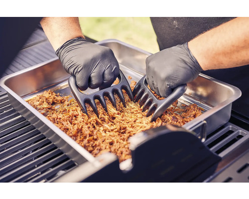 Twee handen met barbecuehandschoenen versnipperen vlees met vleesklauwen in een metalen schaal op een barbecue.