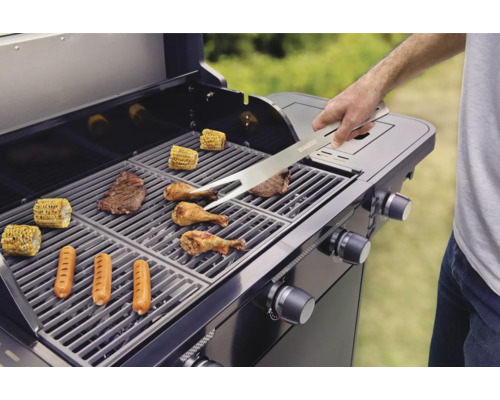 Een barbecue met gegrild voedsel zoals steaks, worstjes, kip en maiskolven wordt met een grilltang omgedraaid.