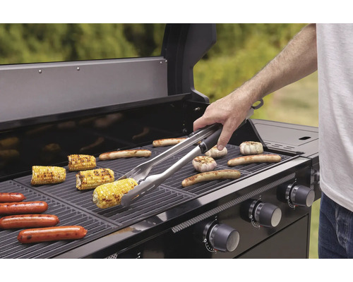 Barbecue met maiskolven, worstjes en knoflook op het rooster en een persoon met een barbecuegereedschap.