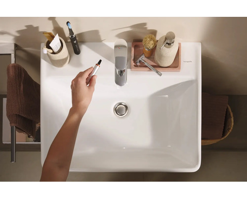 Bovenaanzicht van een badkamerwastafel met kraan en diverse badkamerartikelen, een hand houdt een neushaar trimmer vast.