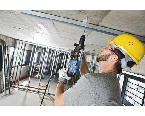 Een man boort met een boormachine in een betonnen plafond, hij draagt een veiligheidshelm.