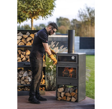Man met veiligheidshandschoenen bereidt een Cook King grilloven in de tuin voor. Houtstapel naast de oven.