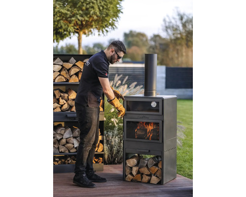 Man met veiligheidshandschoenen bereidt een Cook King grilloven in de tuin voor. Houtstapel naast de oven.