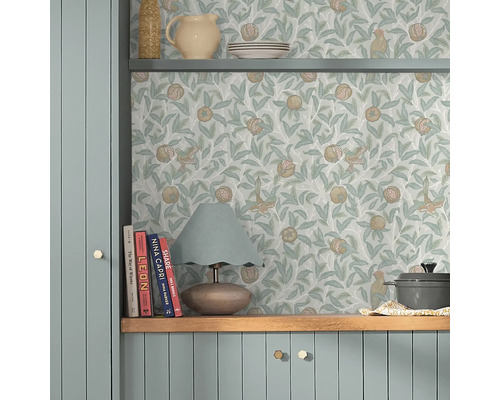 Scene met een plank gedecoreerd met serviesgoed en een lamp, voor een muur met een behang met vogel- en granaatappelmotief.