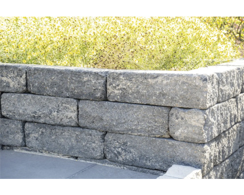 Grijskleurige tuinmuur van stenen blokken met planten op de achtergrond.