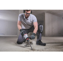 Man knielt en werkt met een boorhamer op een betonnen vloer, terwijl hij beschermende uitrusting draagt.