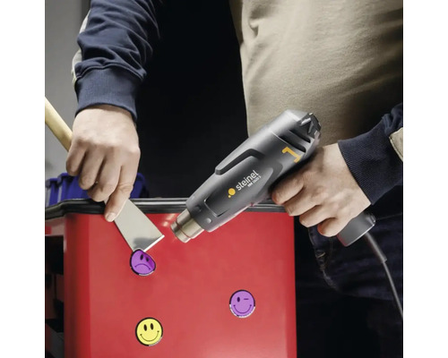 Een persoon verwijdert stickers van een oppervlak met een heteluchtpistool en een plamuurmes.