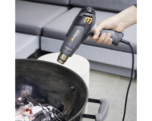 Een persoon steekt met een heteluchtpistool houtskool aan in een barbecue.