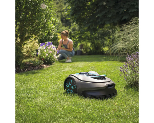 Robotmaaier op gazon met vrouw op de achtergrond die bloemen verzorgt