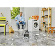 Scène in een bijkeuken met een man, een wasmachine, een rek en een vuilwaterpomp op een betegelde vloer met water