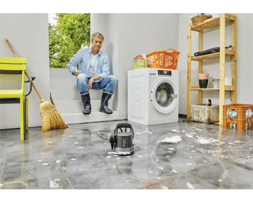 Scène in een bijkeuken met een man, een wasmachine, een rek en een vuilwaterpomp op een betegelde vloer met water