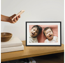 Digitale fotolijst op een houten kast met een foto van twee kinderen die op een roze laken liggen. Een hand houdt er een smartphone boven.