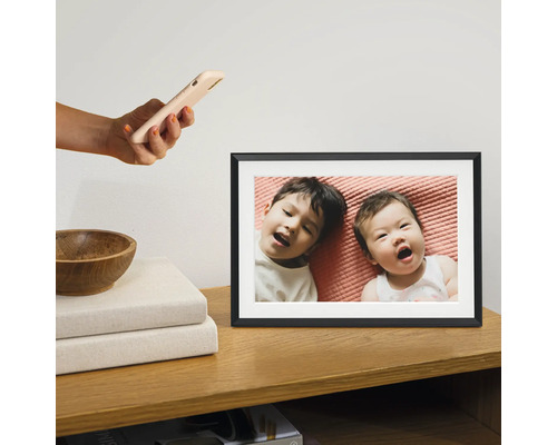 Digitale fotolijst op een houten kast met een foto van twee kinderen die op een roze laken liggen. Een hand houdt er een smartphone boven.