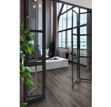Moderne woonkamer met donkere vloer in houtlook, grote industriële ramen en lichte meubels.