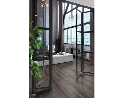 Moderne woonkamer met donkere vloer in houtlook, grote industriële ramen en lichte meubels.