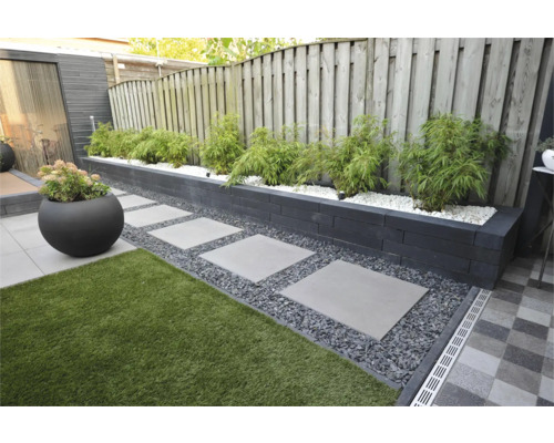Moderne tuin met verhoogde border van donkere stapelblokken, bamboe in wit grind, stapelstenen in donker split en kunstgras.