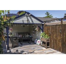 Overdekte zithoek in de tuin met een grijze bank, donkere eettafel, rieten fauteuil en plantenbak op een geplaveid terras.