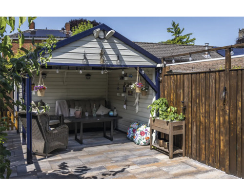 Overdekte zithoek in de tuin met een grijze bank, donkere eettafel, rieten fauteuil en plantenbak op een geplaveid terras.