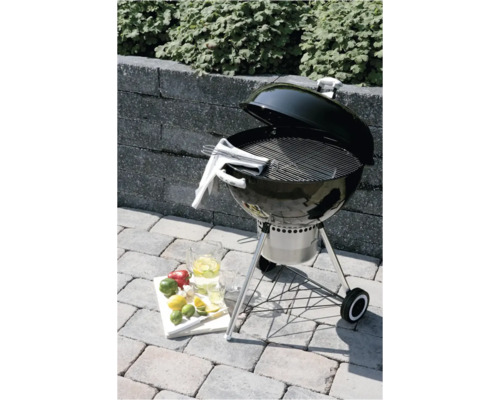 Zwarte houtskoolbarbecue met open deksel op een stenen terras, met roestvrijstalen rooster, asopvangbak en wielen.