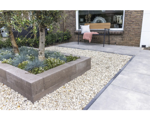 Modern ingerichte tuin met een verhoogde border van grijze stapelblokken, omringd door licht grind en grote terrastegels.