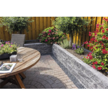 Tuin met een muur van grijze stapelblokken, een ronde houten tafel en diverse bloeiende planten op een bestraat terras.