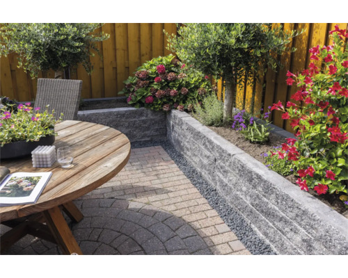 Tuin met een muur van grijze stapelblokken, een ronde houten tafel en diverse bloeiende planten op een bestraat terras.