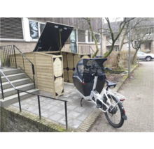 Houten fietsenberging met open deksel en donkere hoekstukken naast een witte bakfiets op een bestrate buitenruimte.