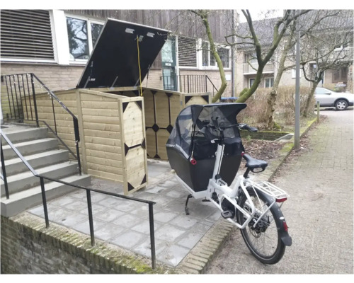 Houten fietsenberging met open deksel en donkere hoekstukken naast een witte bakfiets op een bestrate buitenruimte.