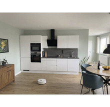 Moderne woonkeuken met witte kasten, grijze achterwand, ronde houten tafel, blauwe stoelen en robotstofzuiger op een lichte houten vloer.