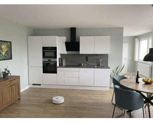 Moderne woonkeuken met witte kasten, grijze achterwand, ronde houten tafel, blauwe stoelen en robotstofzuiger op een lichte houten vloer.