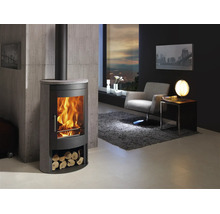 Moderne houtkachel van staal en steen met vuur en houtvak in lichte woonkamer met fauteuil, tapijt en muur in betonlook.