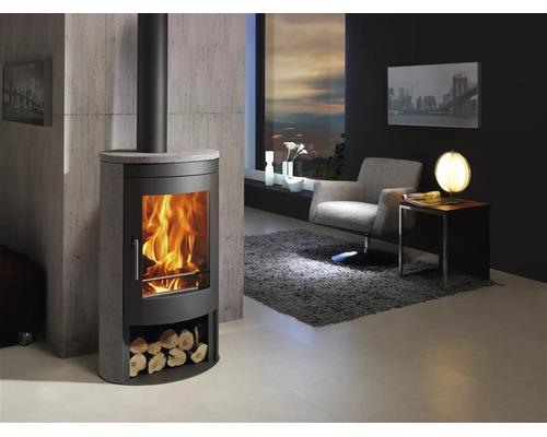 Moderne houtkachel van staal en steen met vuur en houtvak in lichte woonkamer met fauteuil, tapijt en muur in betonlook.