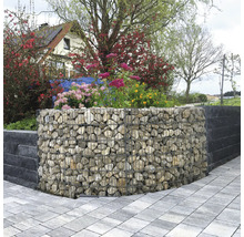 Gebogen schanskorf gevuld met natuursteen als tuinmuur, omgeven door bloeiende planten en een bestraat pad.