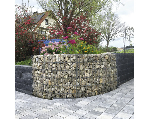 Gebogen schanskorf gevuld met natuursteen als tuinmuur, omgeven door bloeiende planten en een bestraat pad.