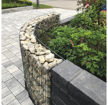 Gebogen schanskorf gevuld met stenen als tuinrand naast een muur en planten op een geplaveide ondergrond.