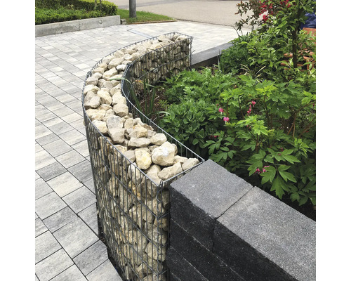 Gebogen schanskorf gevuld met stenen als tuinrand naast een muur en planten op een geplaveide ondergrond.