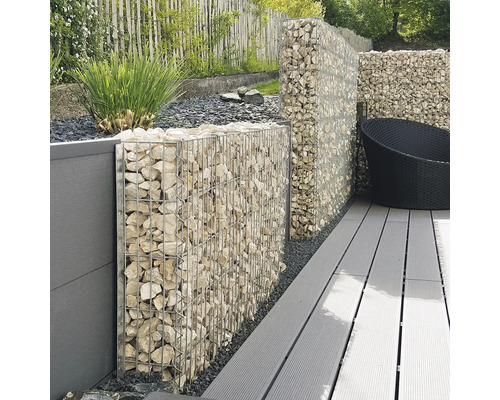 Schanskorf wand gevuld met lichte stenen op een terras met grijze vlonderplanken en een schutting in de tuin.