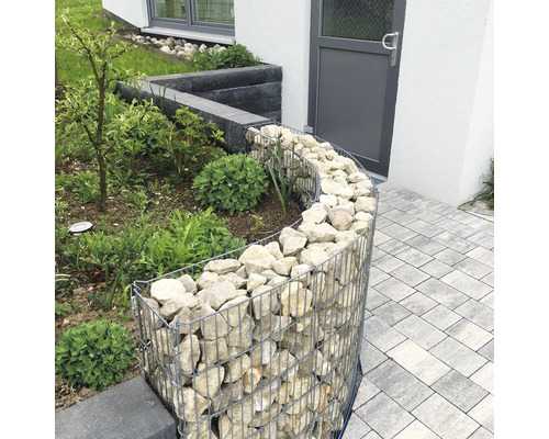 Gebogen schanskorf als tuinborder, gevuld met lichte natuurstenen naast een bestraat pad.