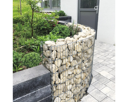 Gebogen schanskorf van gaas gevuld met natuursteen als border voor een tuinperk naast een bestraat pad.