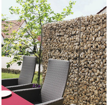 Schanskorf gevuld met natuursteen als schutting in een tuin met loungestoelen van gevlochten kunststof.