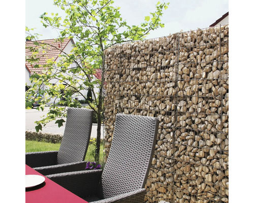Schanskorf gevuld met natuursteen als schutting in een tuin met loungestoelen van gevlochten kunststof.