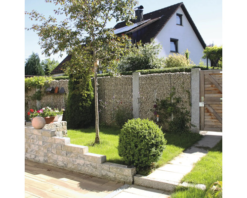 Tuin met een schanskorfwand van natuursteen, granieten palen, houten poort en een lage muur van steenblokken.