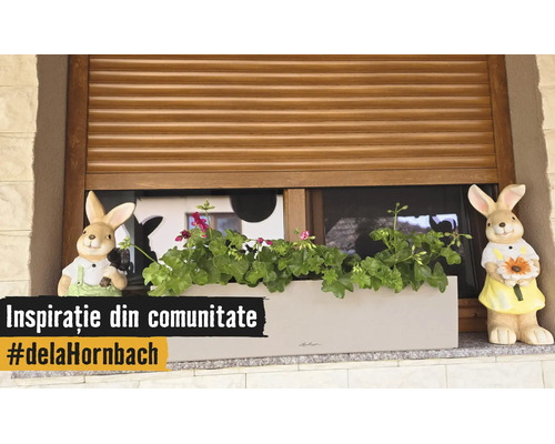 Bloembak met geraniums en twee konijnenfiguren op een vensterbank voor een houten rolluik.