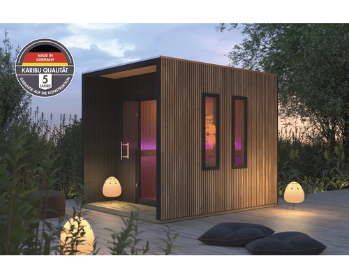 Moderne Karibu tuinsauna met houten lamellengevel en twee ramen, geplaatst op een houten terras met buitenverlichting en zitkussens.
