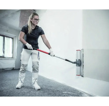 Vrouw gebruikt een breed spackmes met telescoopsteel voor het gladmaken van een muur in een lichte binnenruimte.