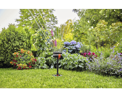 Tuinsproeier in werking op een gazon met bloembedden op de achtergrond.