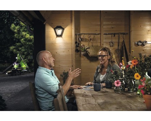 Scene met een man en een vrouw aan een houten tafel voor een houten huis, verlicht door een buitenlamp.