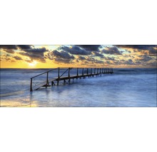 Een houten brug strekt zich uit in de zee onder een bewolkte hemel bij zonsondergang.